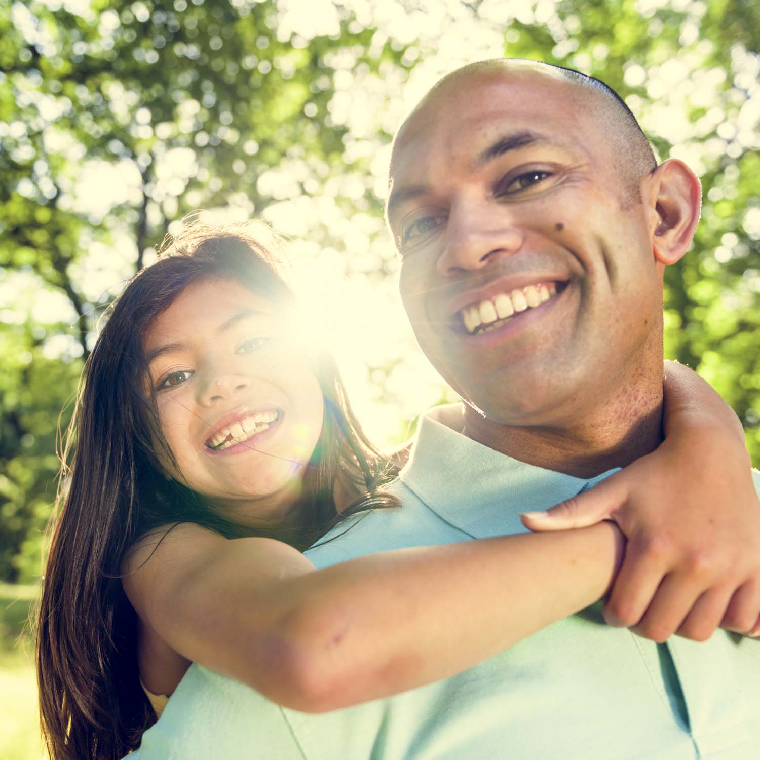 Smiling girl hugging a smiling man from behind under sunlit trees